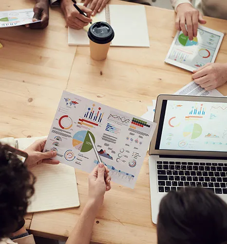 Overhead view of people at a wooden table reviewing printed charts and a laptop showing graphs
