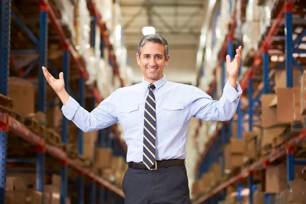 A smiling man in a shirt and tie standing with his arms open in the aisle of a warehouse lined with stocked shelves