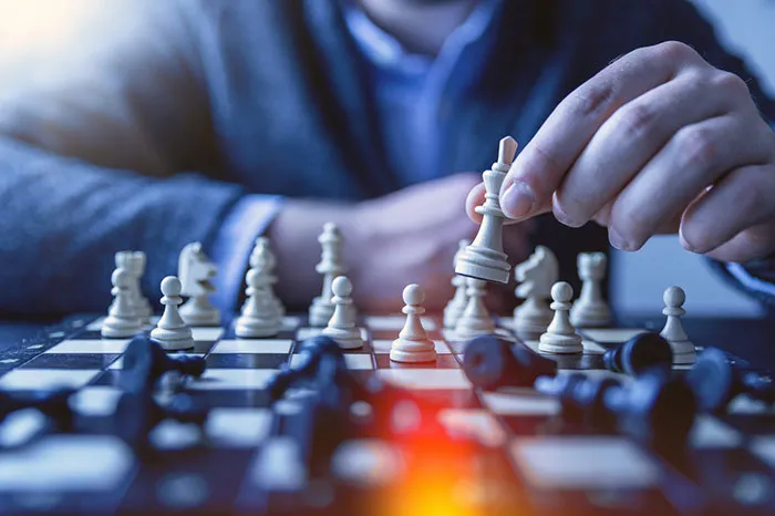 A close-up of a hand moving a white chess piece on a chessboard, with black pieces in the foreground