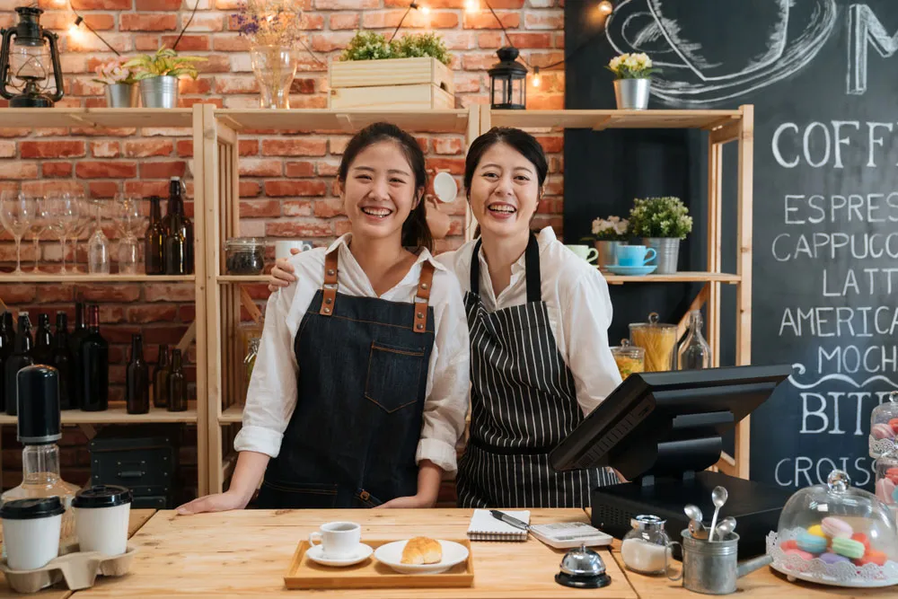 Two smiling cafe owners in aprons behind their counter, with a chalkboard coffee menu on the wall